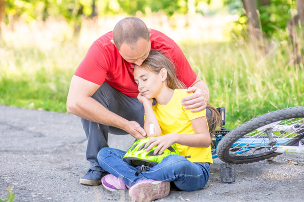 Ein Vater sitzt mit seiner Tochter auf dem Boden, er hält sie sanft und sie schaut entspannt. Neben ihnen liegt ein Fahrrad.